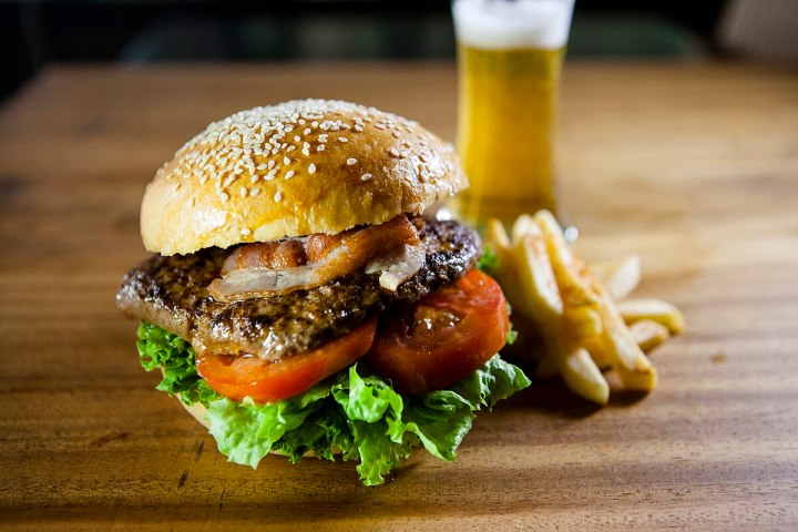 Burger with lettuce, tomato, bacon, fries, and a beer on a wooden table.