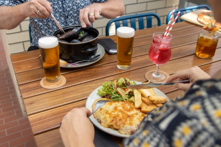 Two people dining outdoors with drinks and plated meals on a wooden table.