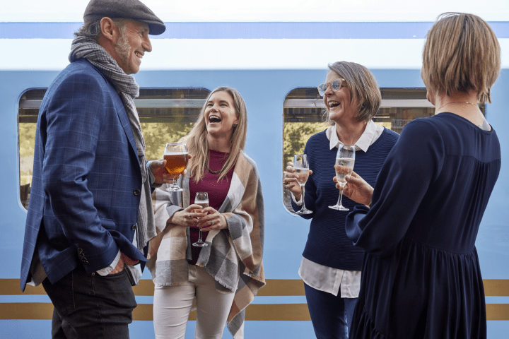 Four people standing by a train laughing and holding drinks.