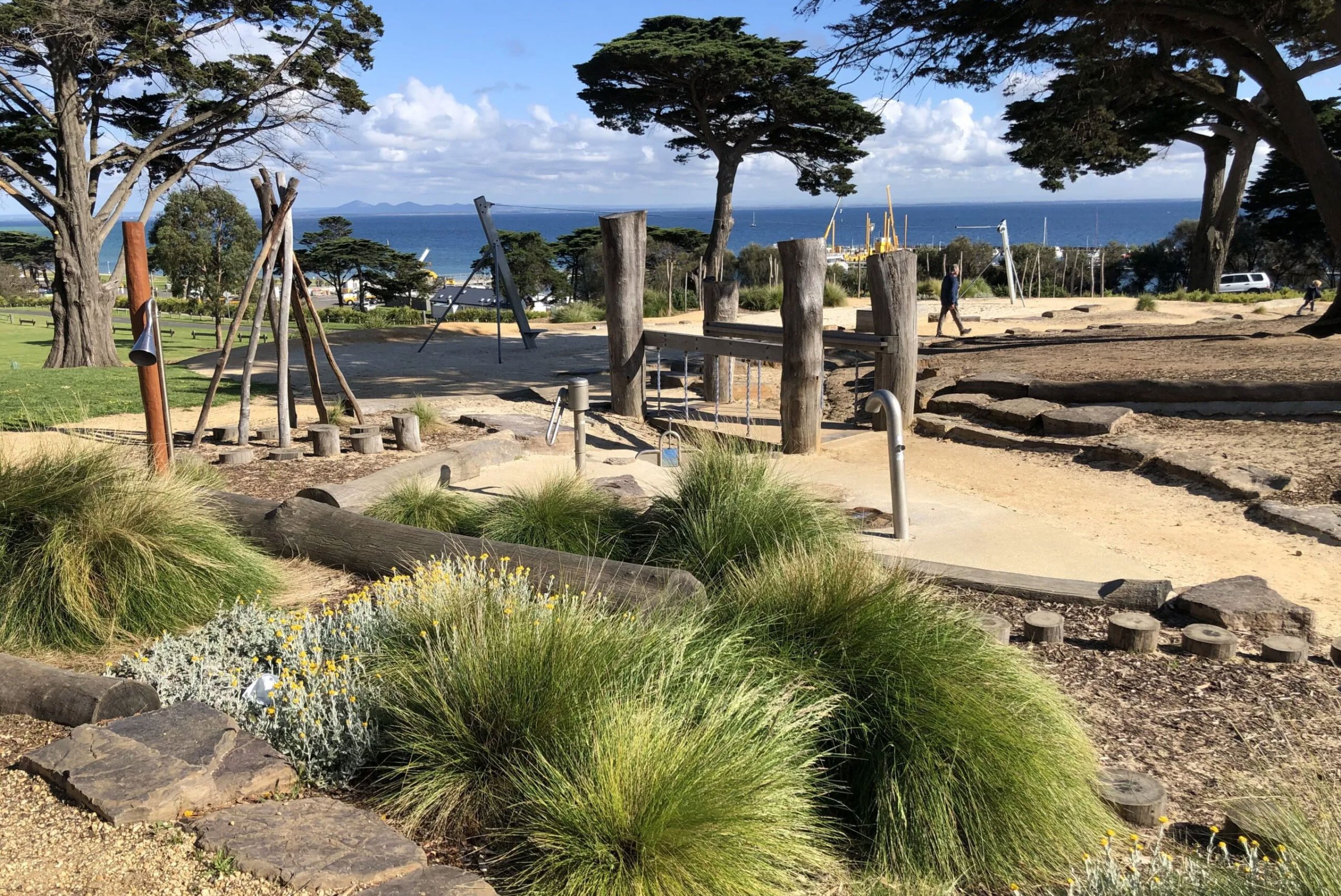 Playground with wooden equipment, sea in background, surrounded by grass and trees.