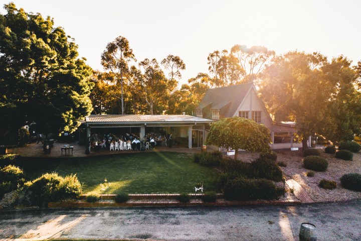 Aerial view of a house with people gathered under a patio in a lush garden setting at sunset.