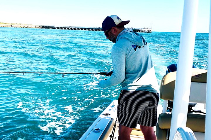 Person in a hat fishing on a boat in clear blue water with a distant pier.