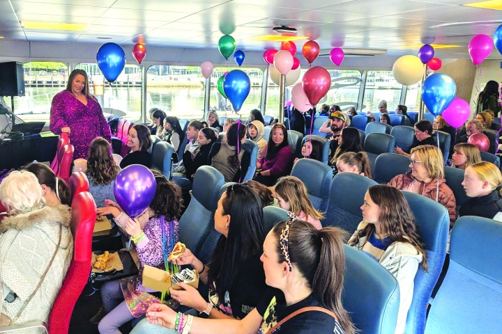 People on a boat celebrate with colorful balloons and food, seated with a view through large windows.