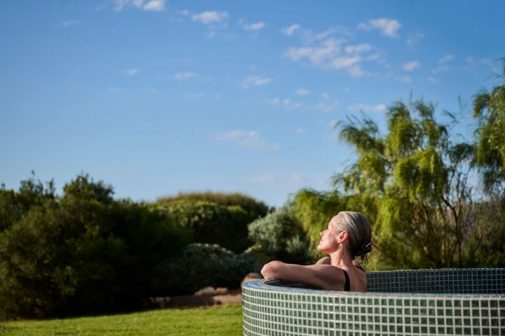 Person relaxing in a tiled outdoor hot tub surrounded by trees and blue sky.