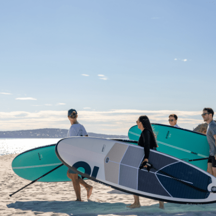 Four people carrying paddleboards on a sunny beach.