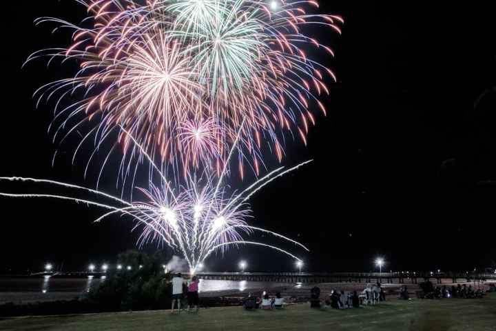 Colorful fireworks over water with people watching on a grassy area at night.