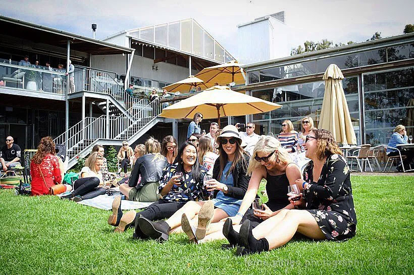 Group of people sitting on grass, enjoying drinks, with a building and umbrellas in the background.