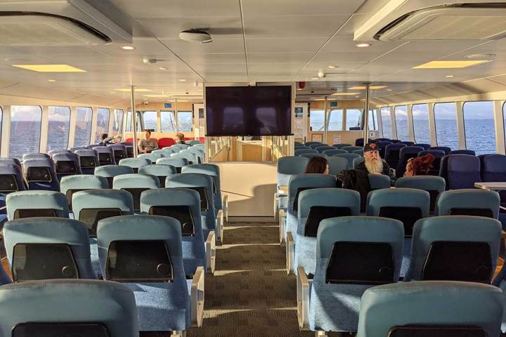 Interior of a ferry with empty blue seats, a screen, and a few passengers sitting near windows.