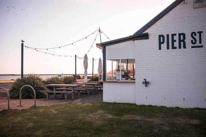Outdoor cafe area with string lights, picnic tables, and white brick building near water.