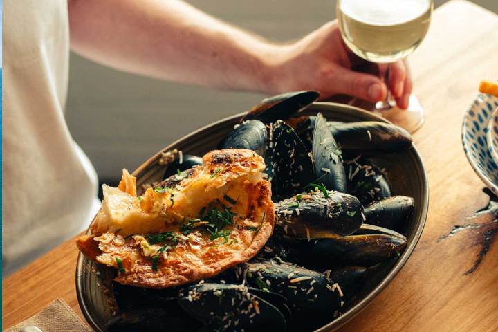 Person holding wine glass, plate of mussels and bread on table.