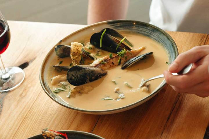 Person eating seafood soup with mussels and herbs, next to a glass of red wine.