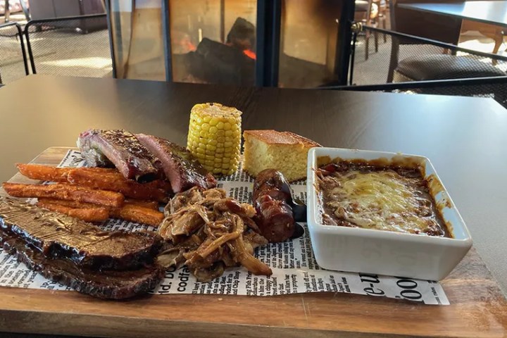 Barbecue platter with meats, cornbread, corn, and beans by a fireplace.