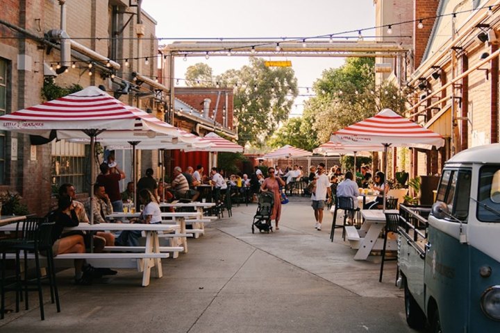 People dining outdoors at tables with striped umbrellas in a sunny, urban setting.