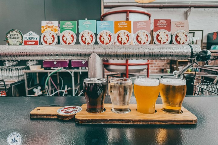 Flight of four craft beers on a wooden tray in a brewery setting.
