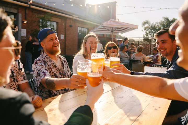 Group of people outdoors clinking glasses of beer at a wooden table.