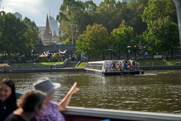 People on a boat pointing toward a riverside park with trees and buildings in the background.