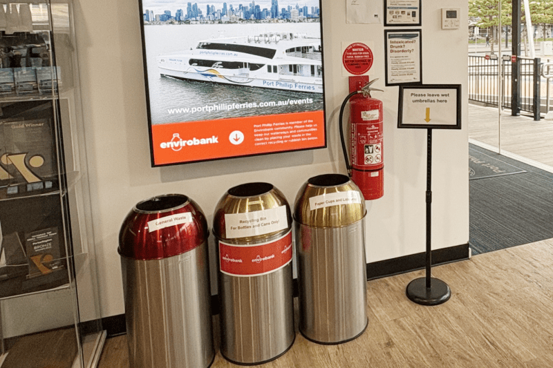 Recycling bins by a wall display, fire extinguisher, and sign indoors.