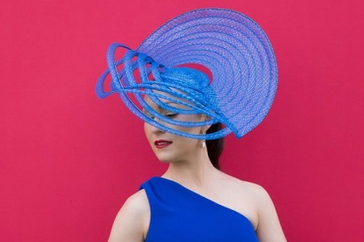 Woman in blue dress and elaborate blue hat against pink background.