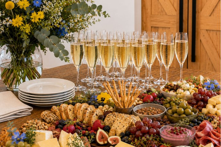 A table with a large charcuterie spread and glasses of champagne, beside a flower vase.